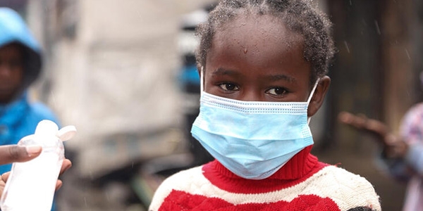 A young girl in Kenya wears a face mask while applying hand sanitizer to help prevent the spread of COVID-19.