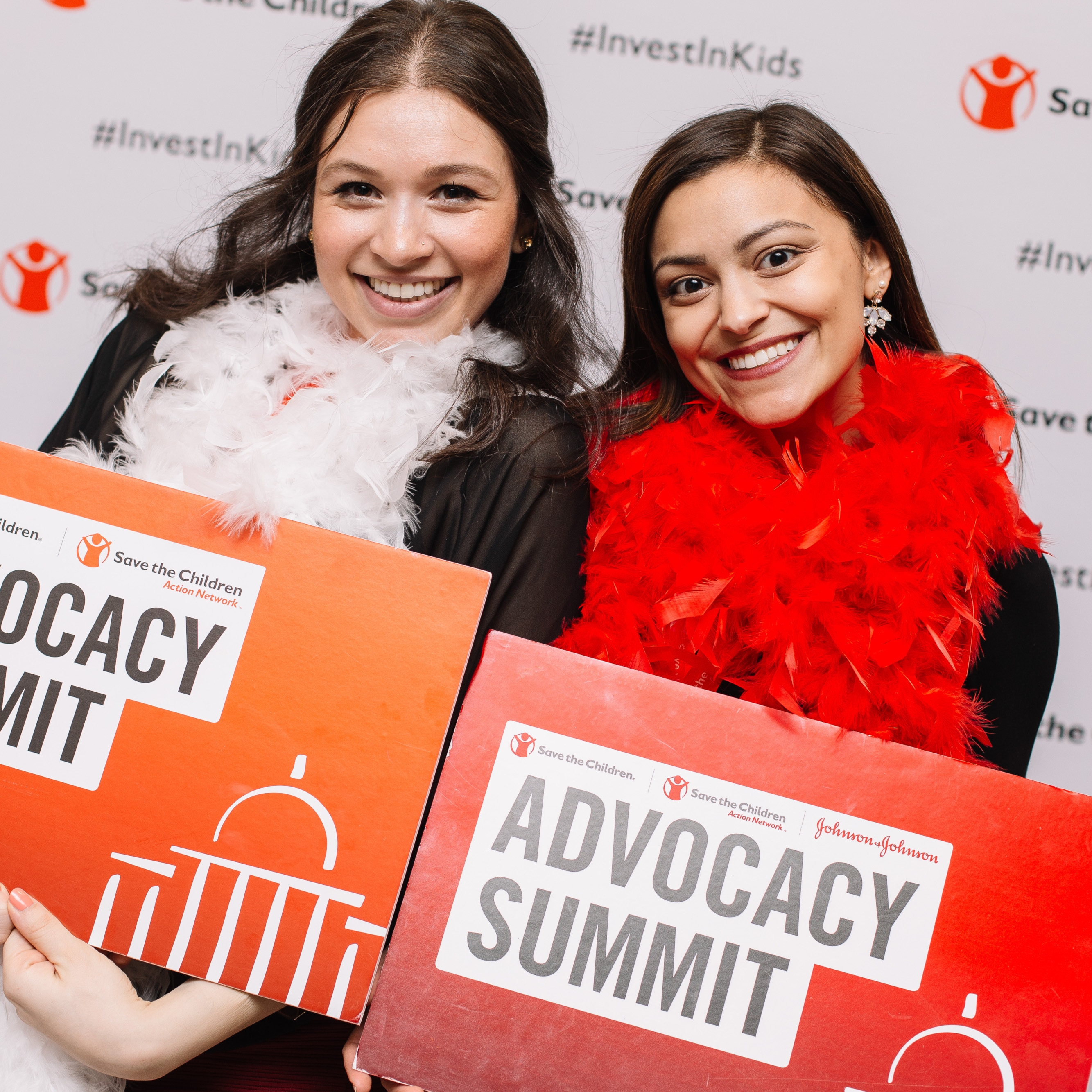 Two Save the Children staff members hold red signs that read, Advocacy Summit: while standing in front of a white mural with Save the Children’s logo and the hashtag #InvestinKids printed on it. Photo credit: Rachel Couch/ Save the Children, March 2019.