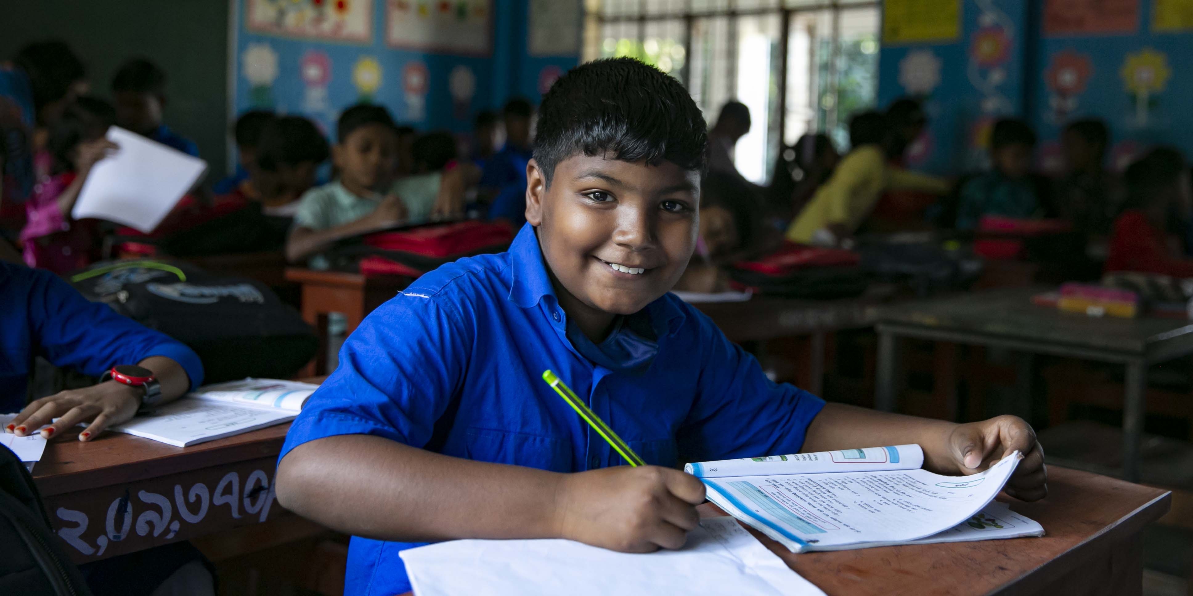 A boy from Bangladesh smiles at the camera while doing school work