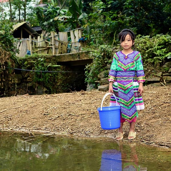Little Tra, 8 years old, wears a colorful outfit stands on the bank of a river near her home, holding a blue plastic bucket.