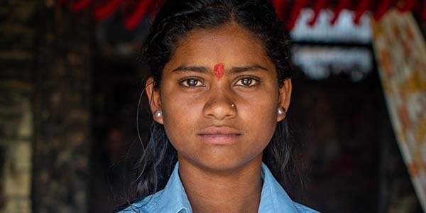 Sixteen-year-old Sonu, wearing her school uniform and a serious expression, holds a tattered textbook in Nepal.