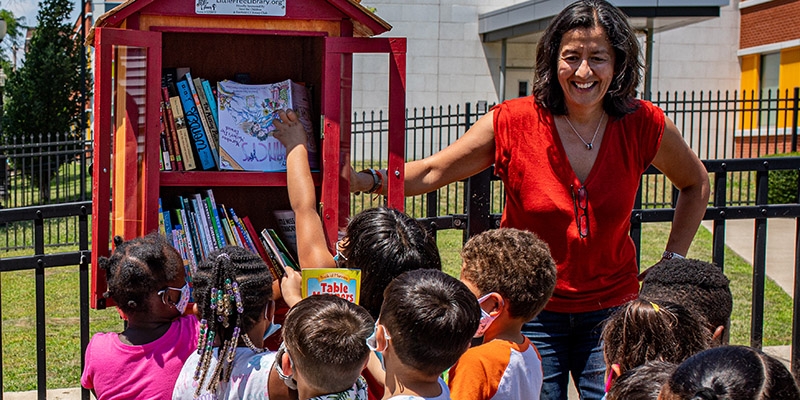 Save the Children CEO Janti Soeripto stands near a Little Free Library while children enjoy book selection. 