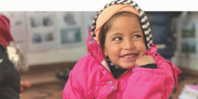 A little girl in a pink jacket smiles while looking away from the camera.