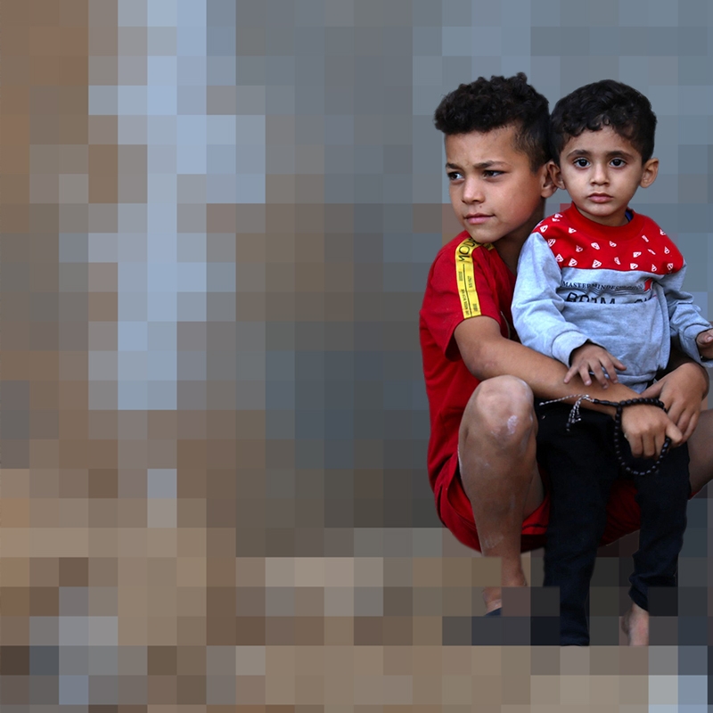 An image with a blurred background shows two boys sitting together in an area destroyed by conflict. 