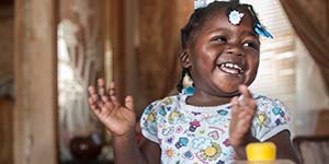 A 20-month old girl who lives in Mississippi smiles as she gets ready to clap her hands while standing near a table with a bright toy on it. 