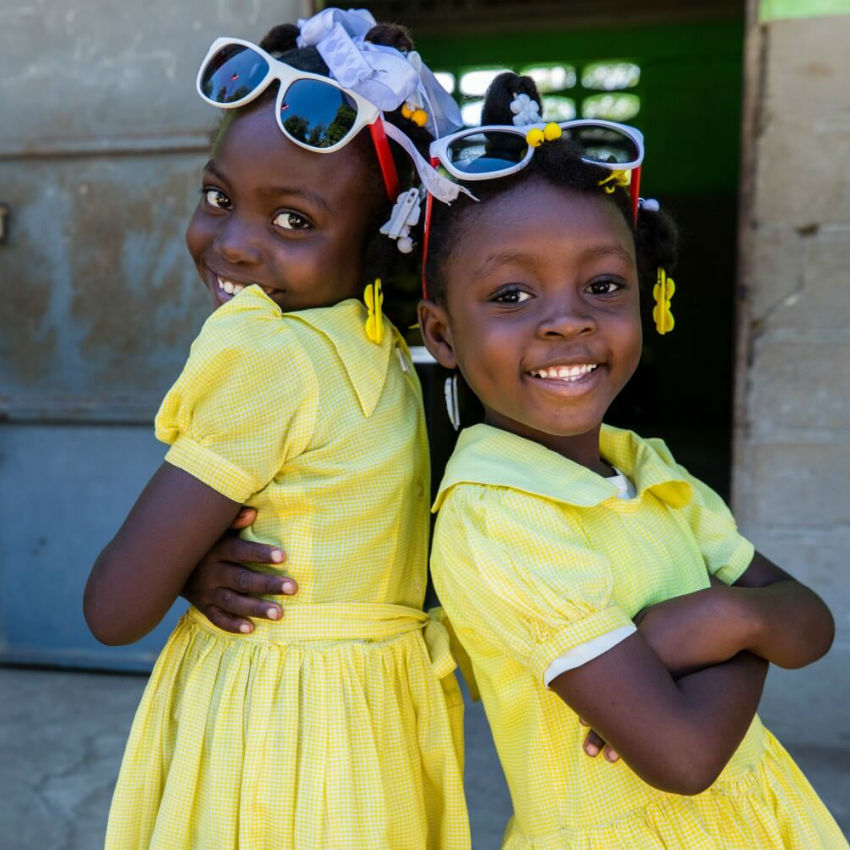 In Haiti, two girls wear yellow school uniforms and sunglasses while standing back-to-back with one another and smiling. 