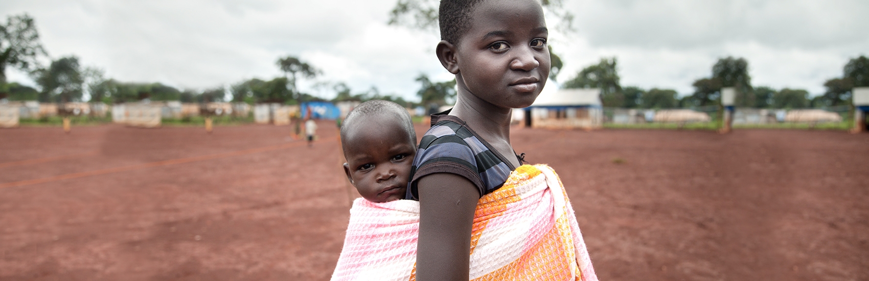 In Burundi, a girl holds a baby on her back in a fabric sling while standing near homes in her village. 