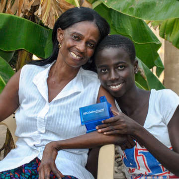 An adolescent girl from Ghana, stands proudly next to her mother as she displays a small, blue plastic bank from the YouthSave program.  She saves about a $1 a week through a savings account provided by YouthSave’s partner in Ghana, HFC Bank. Photo Credit: Adadzewa Otoo/Save the Children 2013.