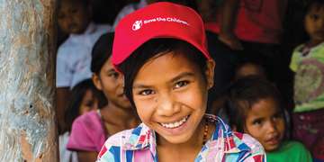 An 11-year-old Indonesian girl, wearing a Save the Children cap, smiles for the camera. She wasn’t taught to speak the local language (Bahasa Indonesia) properly, due to her parents’ lack of education; this affects her ability to express herself to her peers and teachers. She attends a Save the Children education program in West Sumba, Indonesia to get the extra help she needs. Photo credit: Victoria Zegler/Save the Children, May 2017. 