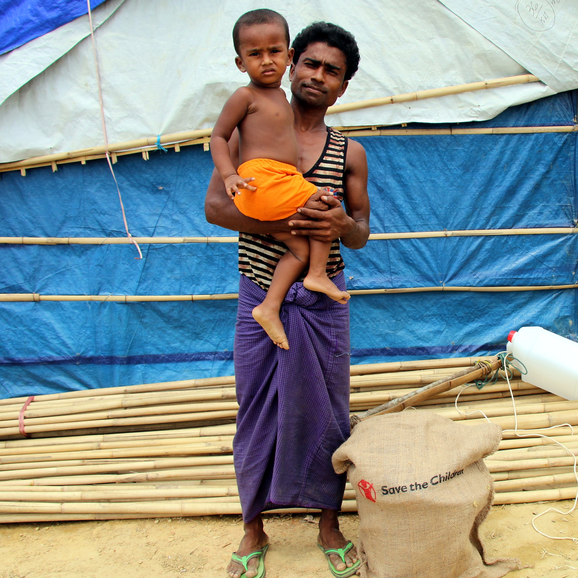 A father holds his two-year-old son in front of a makeshift hut in a Rohingya refugee camp in Cox’s Bazar, Bangladesh. He and his wife fled  Myanmar in August 2017, when violence escalated. The family walked for seven days to get to Bangladesh, and when they arrived, the child was extremely sick with diarrhea. Photo credit: Daphnee Cook / Save the Children, May 2018. 