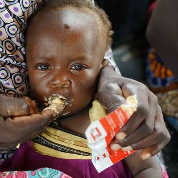 An infant of 9 months, with her mother Alima, eats high nutrient peanut paste as a distribution at the Guidanaoutchi Clinic, Niger. Photo Credit Save the Children 2016.