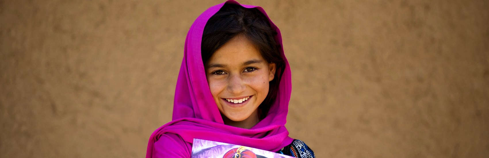 9-year old Najma holds a book and smiles outside her home in Pakistan. Najma participates in a reading “buddy program,” where she helps another child with reading in and outside of school. The buddy program is part of Save the Children’s Literacy Boost initiative, which includes assessments, teacher training and community action, to significantly improve children’s core reading skills. Photo credit: Asad Zaidi/Save the Children, January 2015. 