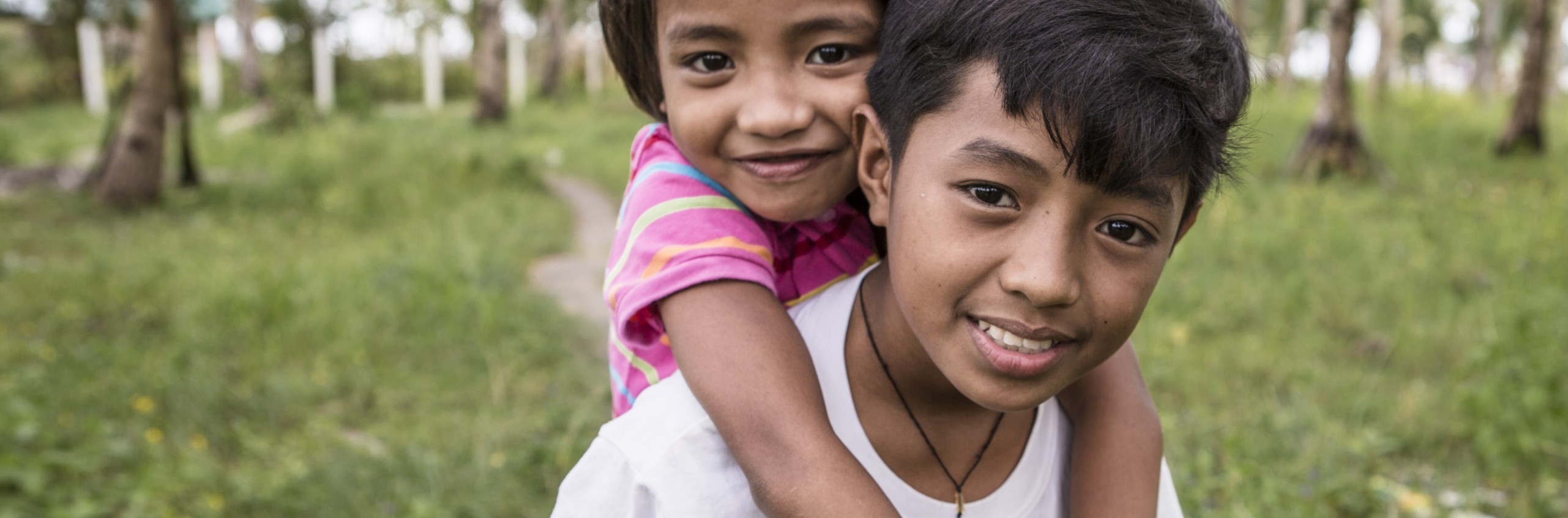 Philippines, a little girl in a pink shirt smiling, is on the back of a little boy in a white t-shirt who is also smiling at the camera.
