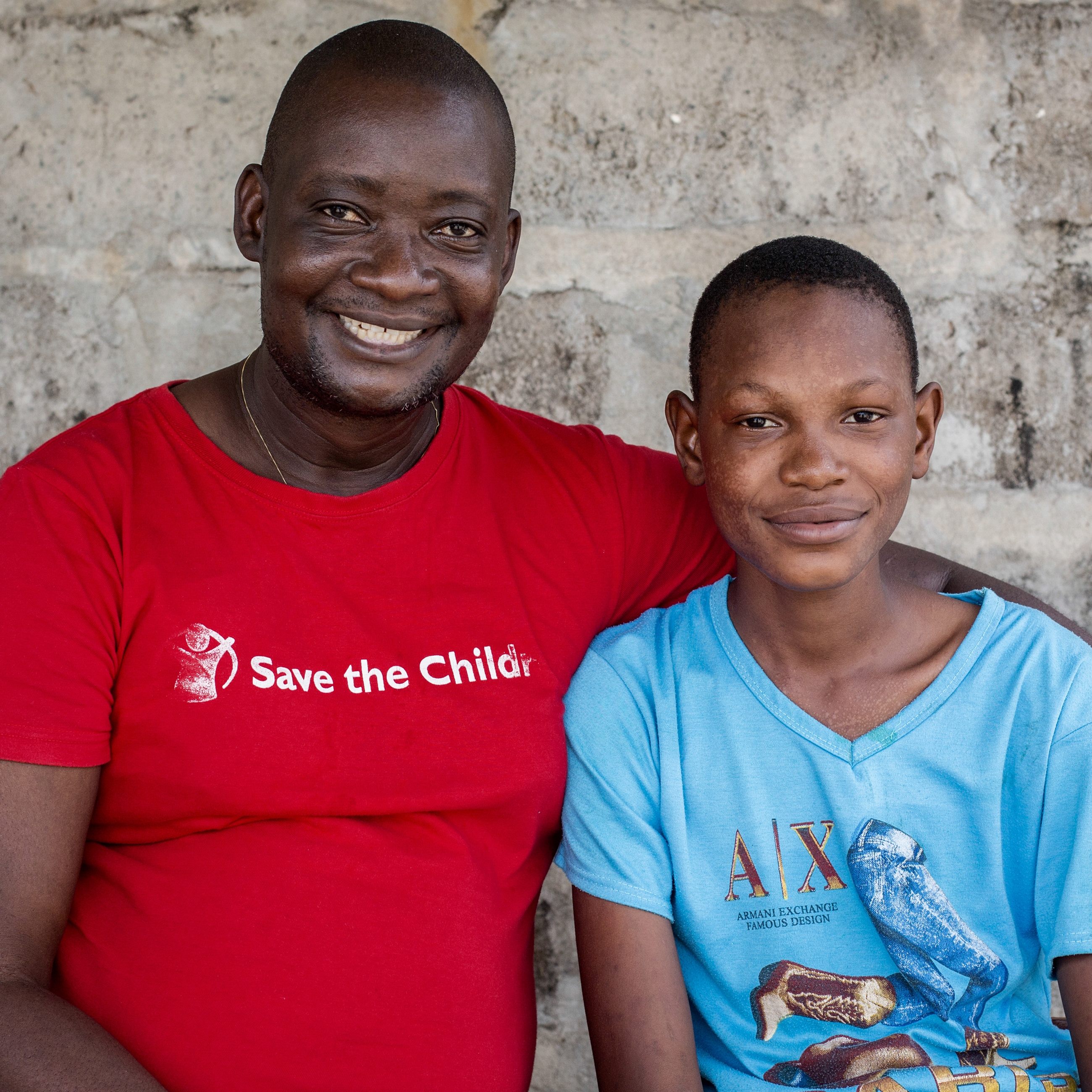Child protection officer, Konday Marah, and 16-year-old Ebola survivor Joshua* sit outside a Save the Children field office in Sierra Leone. Thanks to the regular visits with Konday and our work to help reintegrate children who have survived Ebola back into society, Joshua’s life is returning to that of a typical 16-year-old boy. Photo Credit: Jonathan Hyams/Save the Children 2015.