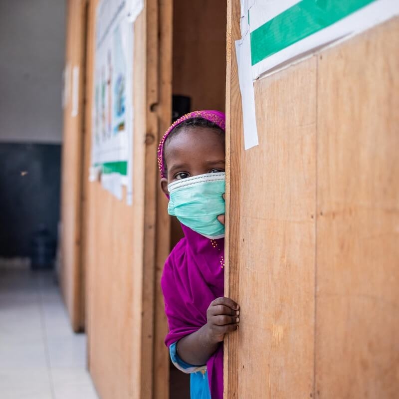 A child wearing a face mask peers out from behind the wall of a health clinic in Somalia.