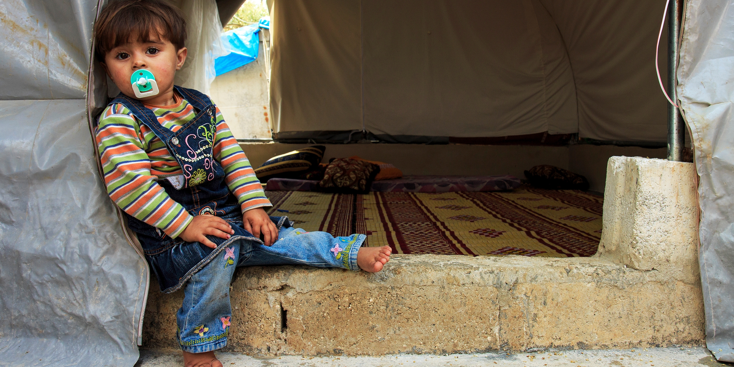 A two-year-old girl sits on the floor of her family’s tent in a camp for displaced people on the northern Syrian border. Photo credit: Ahmad Baroudi/Save the Children, May 2015. 