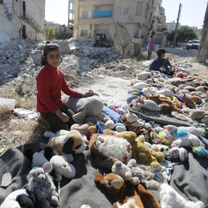 A little girl sits along a street in Syria lined with stuffed animals on the ground. There are two other kids in the background.
