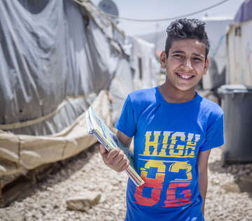 A 13-year old boy stands with his school books outdoors in an informal tented settlement in Lebanon.
