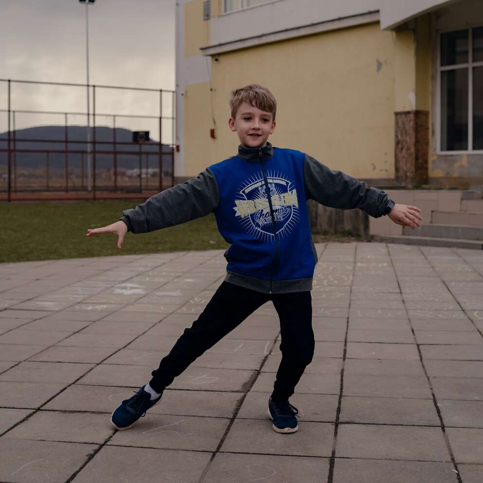 In Ukraine a boy stands outside on a paved area near a yellow building. 