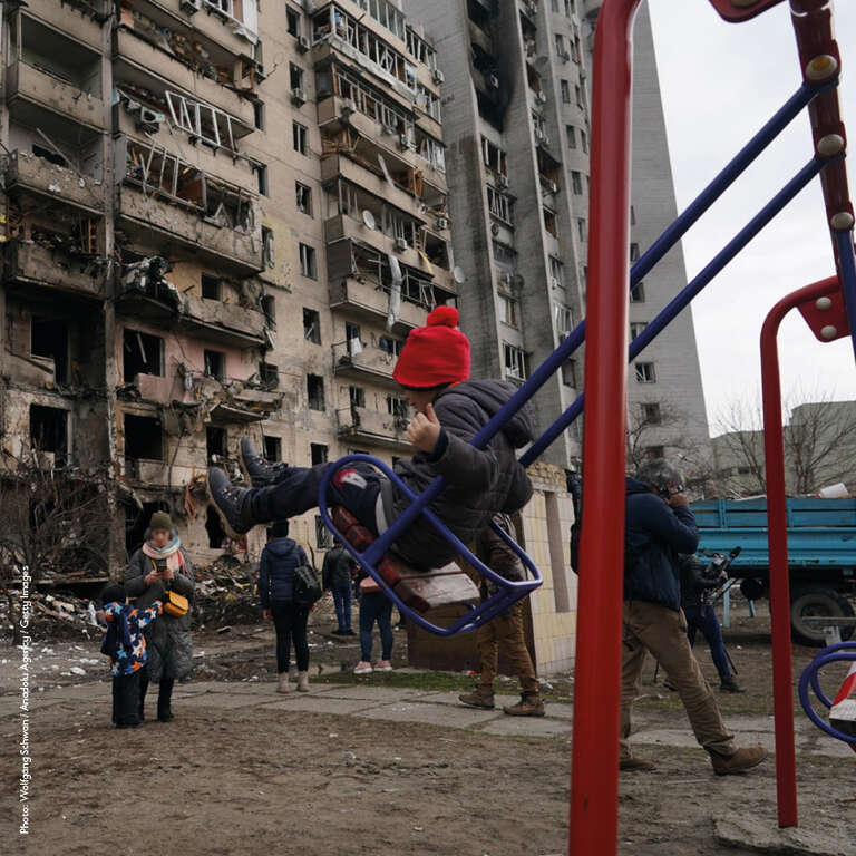 In Ukraine, a child swings on a playground swing near a building that has been destroyed in a missle attack. 