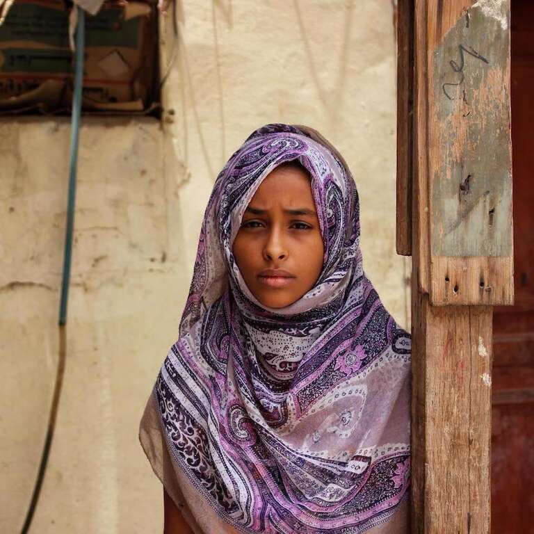 A girl stands in front of a wall marked with bullet holes in Yemen.