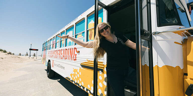 A smiling woman leans out of a bus with her arm outstretched. The bus is wrapped with a 100 Days of Reading logo.