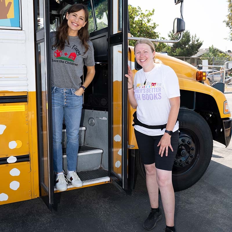 Save the Children Ambassador Jennifer Garner stands in the doorway of a yellow school bus and next to a woman wearing a Summer's Better with Books t-shirt.
