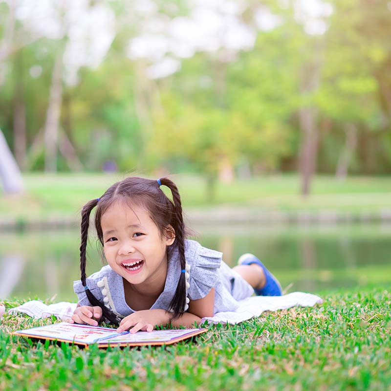 A young girl lays on the grass in a park and enjoys reading from an open picture book.