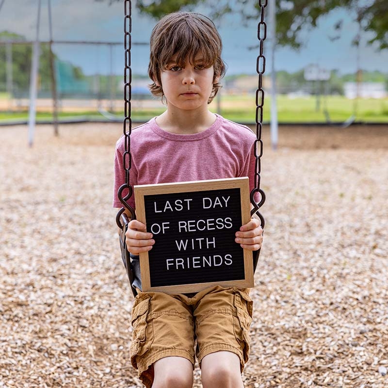 A student sits on a swing in an empty playground and holds s sign that reads, Last Day of Reading. 