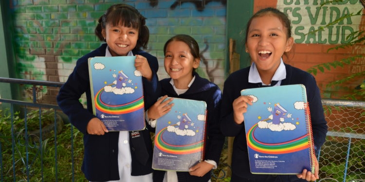 A group of three girls wearing school uniforms hold books outside a school in Bolivia.
