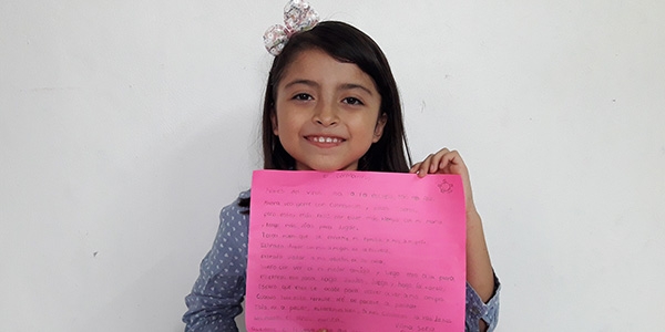A smiling girl holds up a poem written on red construction paper. 