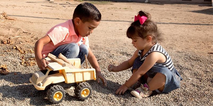 A young boy and girl play with a yellow toy truck in the dirt outside a home in rural America.