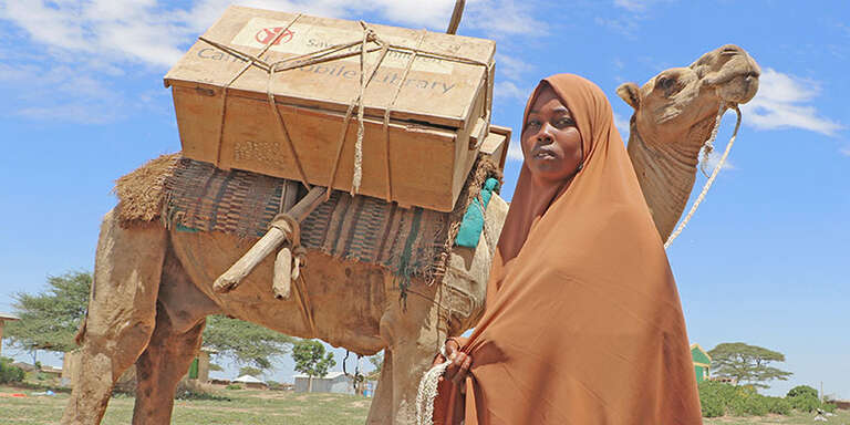 A girl stands next to a camel carrying a mobile library.