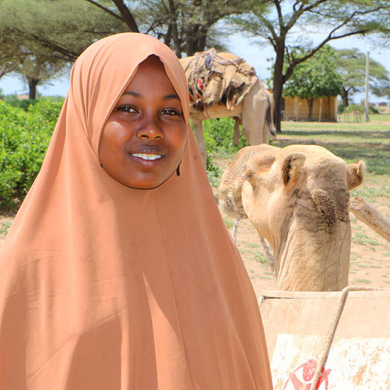 13-year-old Maha stands beside a camel, wearing a peach-colored headscarf. She’s squinting in the bright sun and smiling into the camera. Beside her is a camel with a large box strapped on its back, part of a Save the Children Camel Library initiative. Camels traverse the desert, bringing books to children who are out of school due to the pandemic. Photo credit: Seifu Asseged / Save the Children, September 2020. 