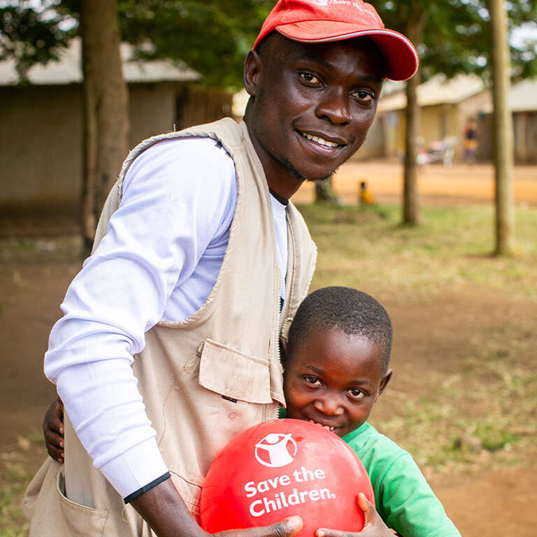 Provy hugging a Save the Children worker. The both hold a red balloon with the Save the Children logo. 
