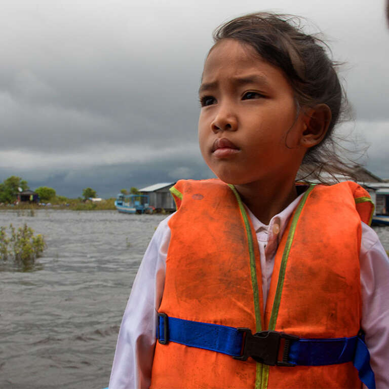 Seima wearing an orange life jacket, with a cloudy sky and water in the background. 