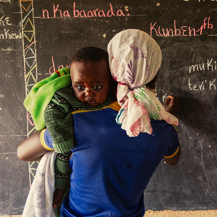 A young mother holds her son, while she writes on a blackboard at a Save the Children learning and livelihood center.