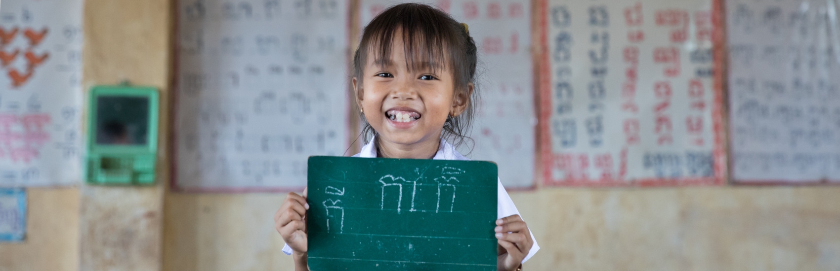 Little Monyrath, age 7, one of our First Read program students, stands at the front of her classroom, proudly holding a mini chalkboard displaying her work.