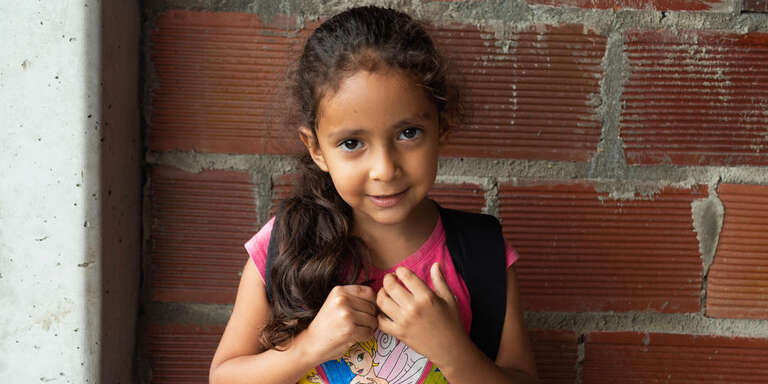 A young girl wearing a back pack with black straps stands in front of a red brick wall.