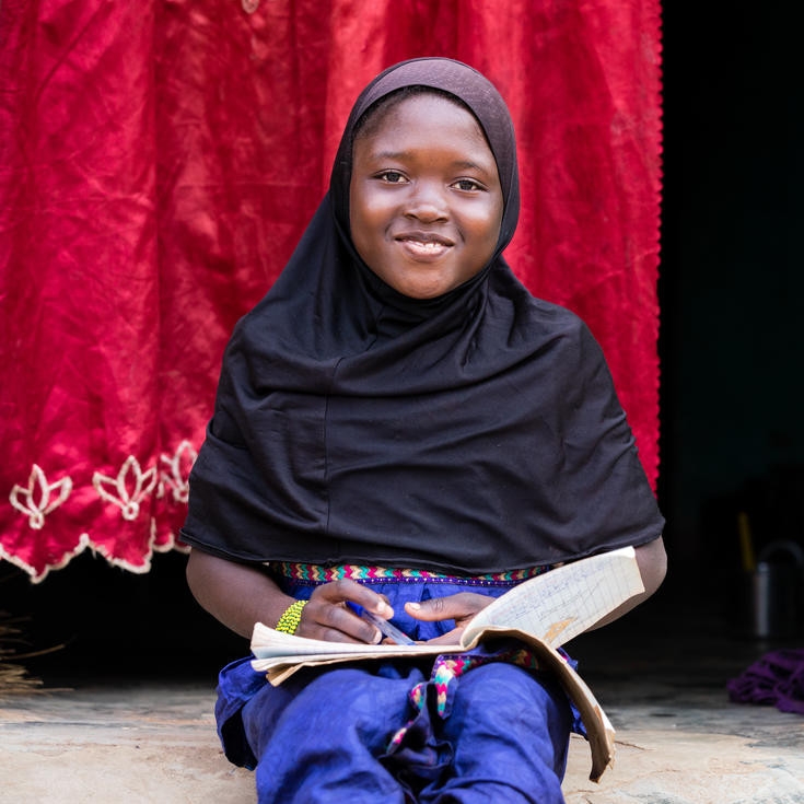 Awa, age 10, sits and smiles in her home’s doorway, covered by a bright red curtain, while working on her homework.