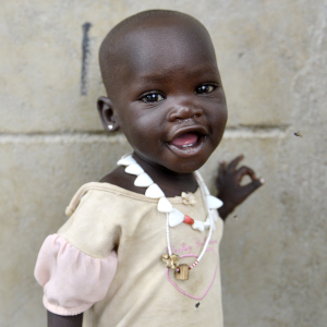 A one-year-old smiles as she places one hand on a wall near a food distribution center in South Sudan.