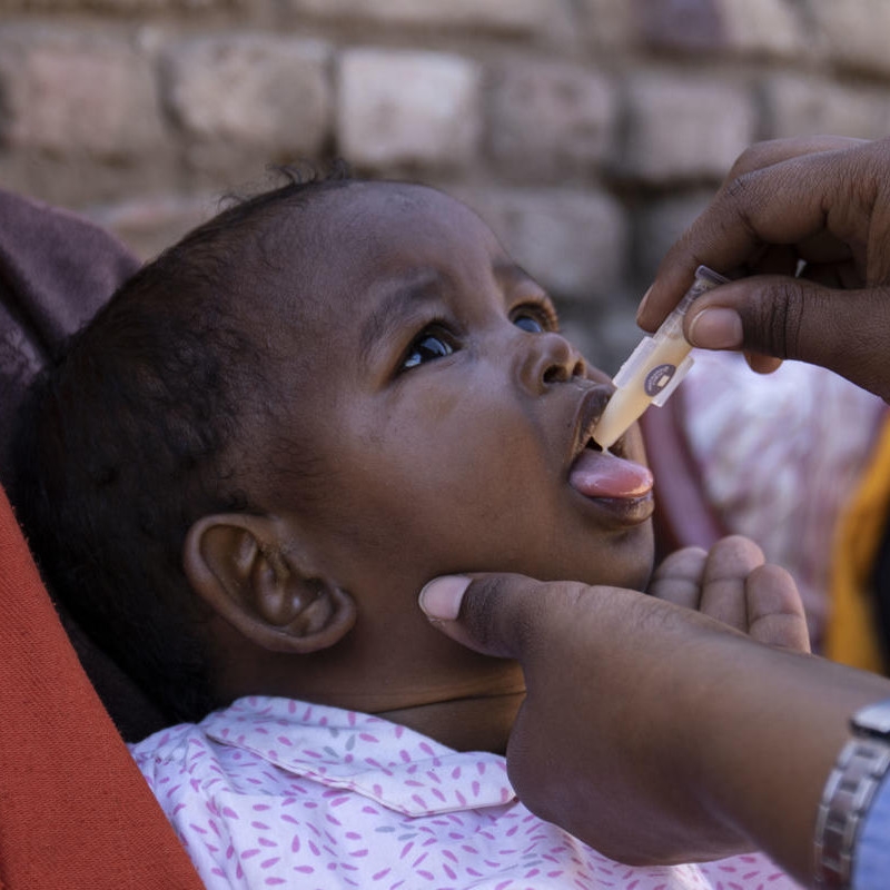 Little 1-year-old Manar, sitting in her mother’s lap, opens her mouth to receive an oral cholera vaccine from a Save the Children health worker.