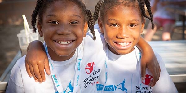 Two girls wearing Save the Children shirts smile at the camera. 
