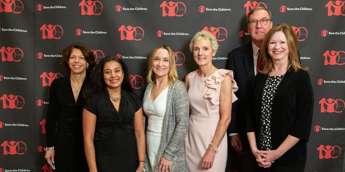 Six member of Save the Children’s Boston Leadership Council Benefit pose in front of a patterned background printed with the Save the Children logo. The men and women are attended a benefit at the Downtown Harvard Club hosted by Tim Douglas. Photo credit: Casey Atkins/Nov 2018.