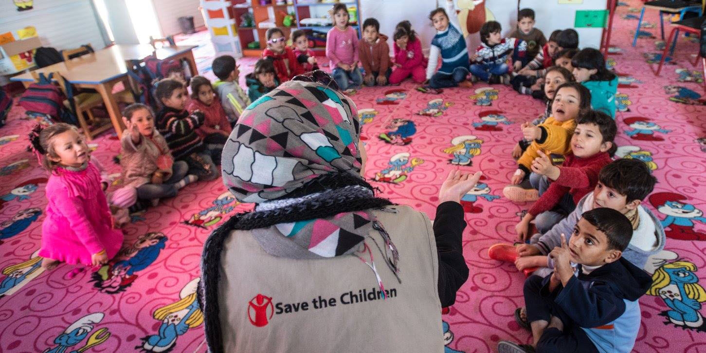 A woman wearing a Save the Children branded shirt sits with a group of children in a circle.