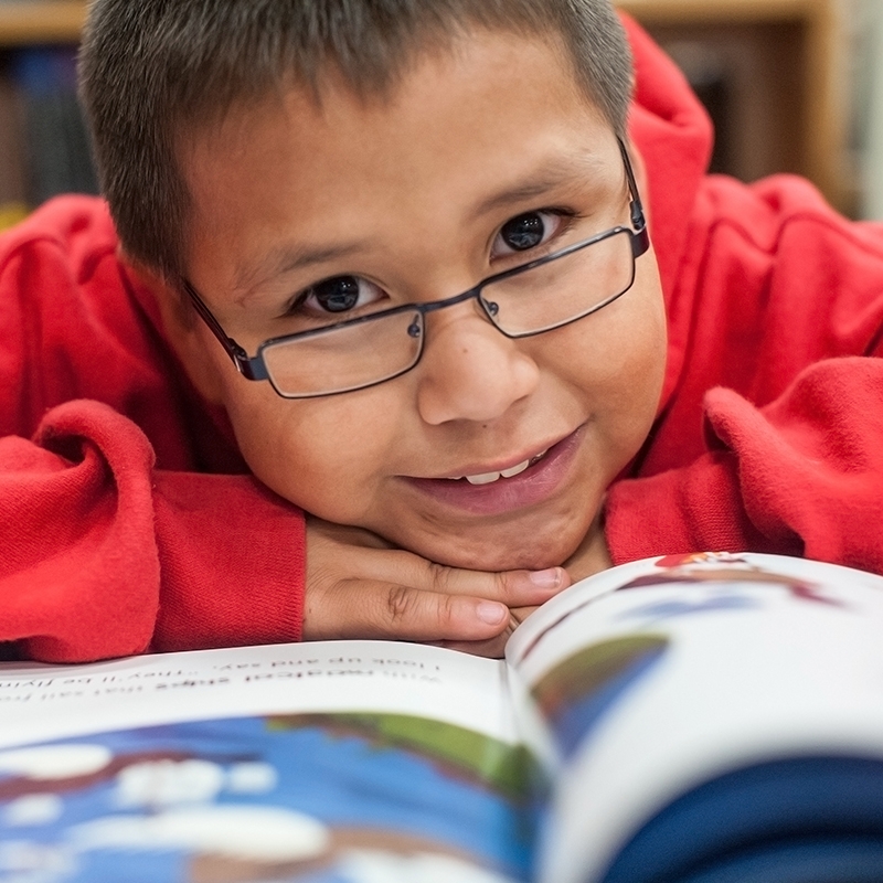A young boy wears a red sweatshirt while resting his head on an open book.