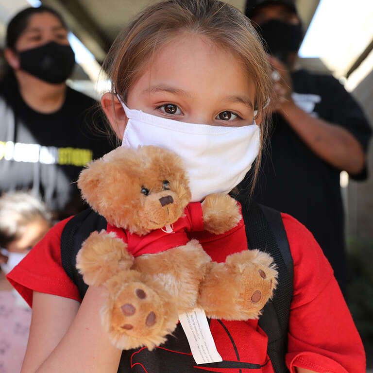 A girl in a face mask holds up a teddy bear.