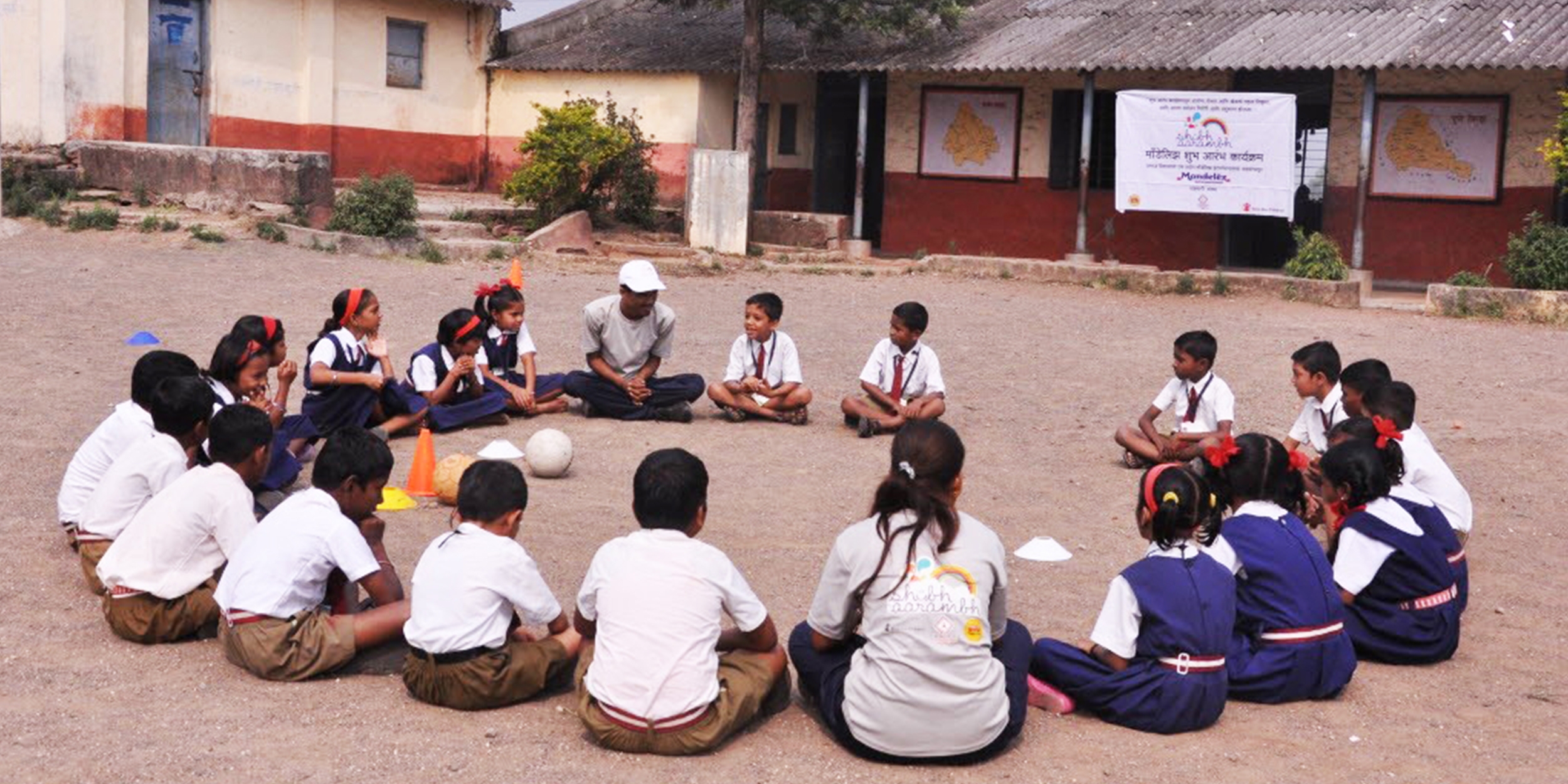 A group of children sit on the ground in a circle at a school in Induri, Maharashtra, India. Their attention is on a teacher. These play sessions are sponsored by Mondelez, and involve learning nutrition and playing games to reinforce a positive message about exercise and good nutrition. Photo credit: Luciana Bonifacio / Save the Children, November 2015. 