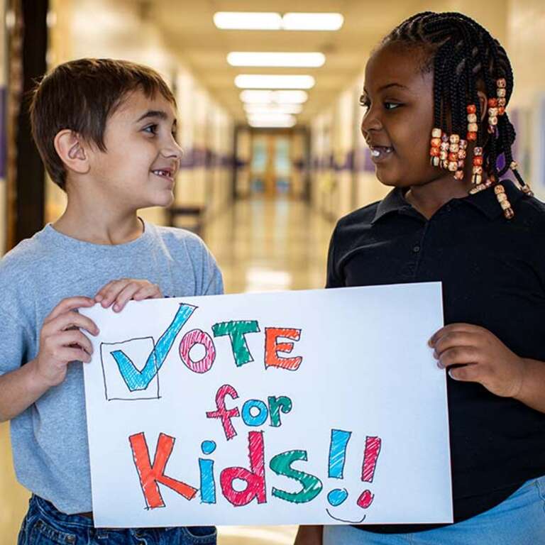 A boy and girl hold a sign that says "Vote for Kids."