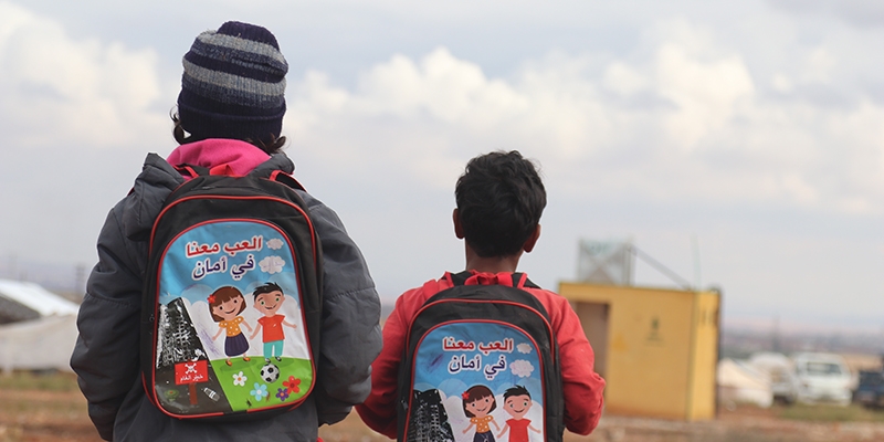Two children wearing backpacks walk away from the camera. 